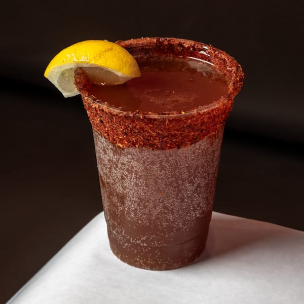 A michelada in a plastic cup with a chili-lime rim and a lemon wedge.