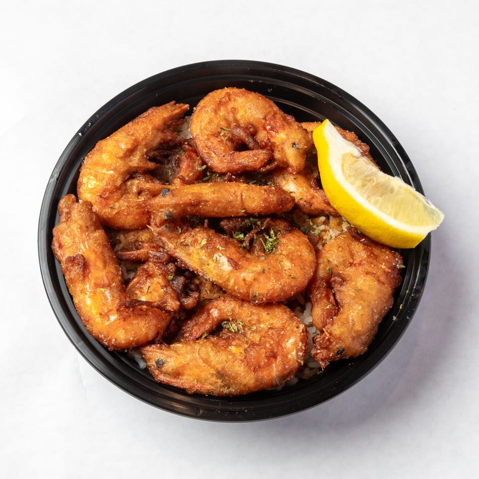 Crispy fried shrimp with herbs and a lemon wedge in a black bowl on a white surface.