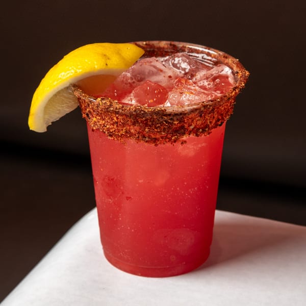 A vibrant red drink with ice, garnished with a lemon wedge and a chili-lime rim.
