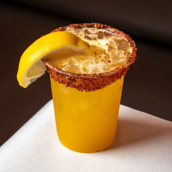 A vibrant yellow drink with ice, garnished with a lemon wedge and a chili-salt rim.