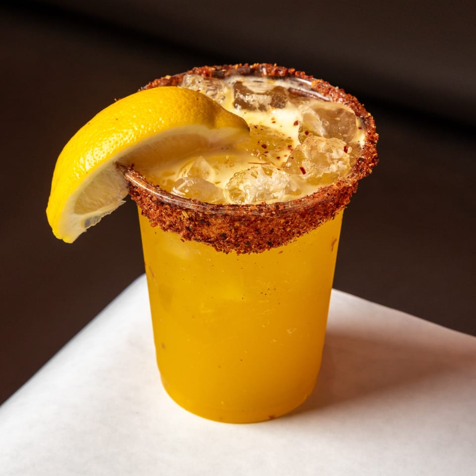 A vibrant yellow drink with ice, garnished with a lemon wedge and a chili-salt rim.